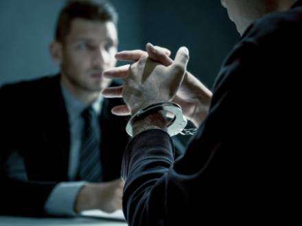 A person in handcuffs sits at a table, with another individual in a suit facing them, engaged in conversation.
